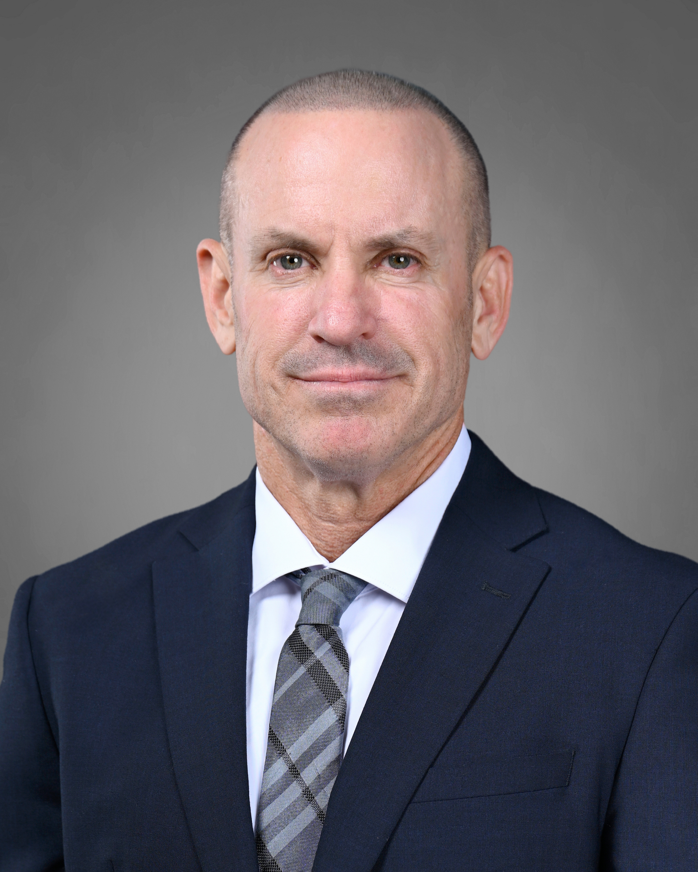 Professional headshot of a man in a navy suit, white shirt, and gray plaid tie against a gray background.